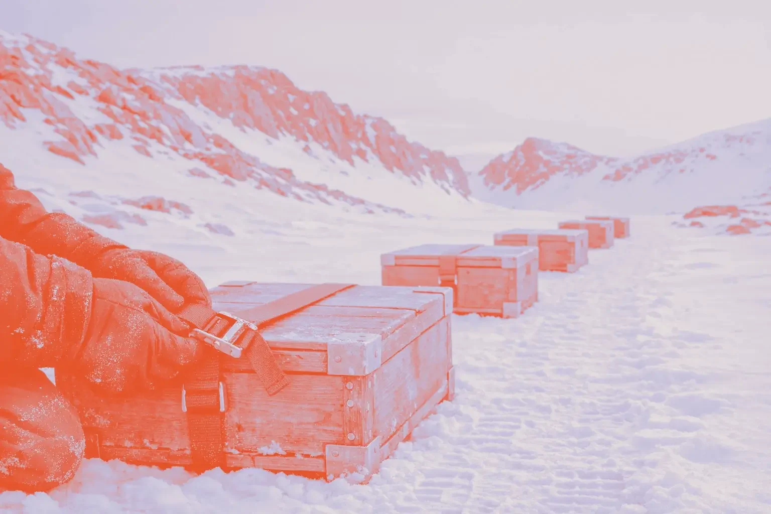 A person fastens a green strap on a wooden box in a snowy landscape with several other boxes in different colors lined up behind.