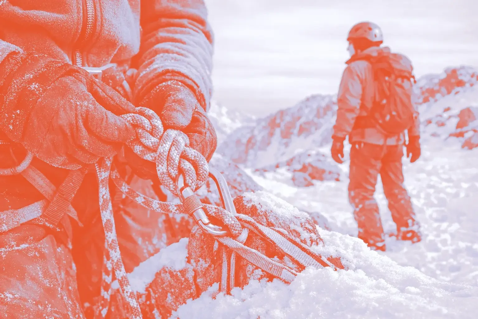 A close-up of a person's hands tying a knot with climbing rope, with a person in red standing in the background on snow-covered rocks.