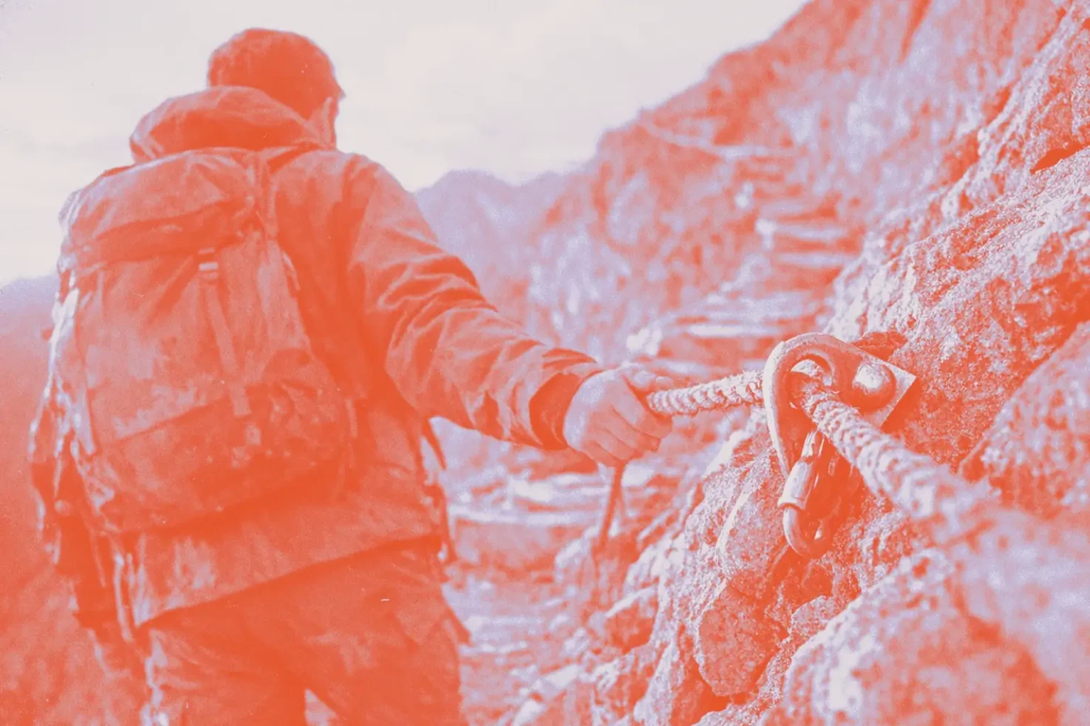 A backpacked climber seen from behind reaching for a steel via ferrata cable anchored to rock on an exposed mountain route, with switchback terrain below.