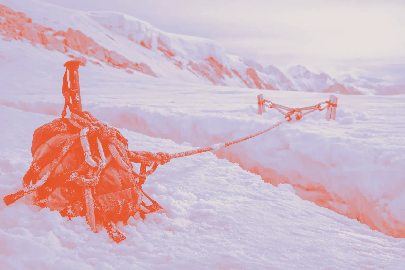 A black backpack with climbing equipment rests on a snowy surface beside a crevice.