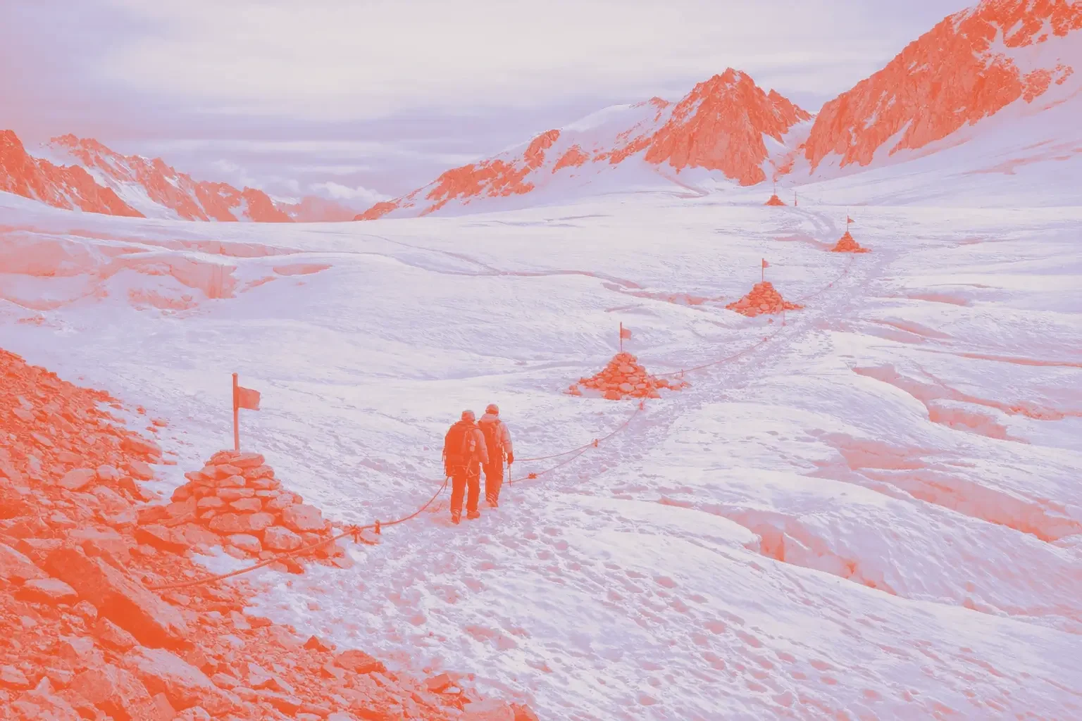 Two hikers walk on a snow-covered glacier, marked by red flags and stone cairns.
