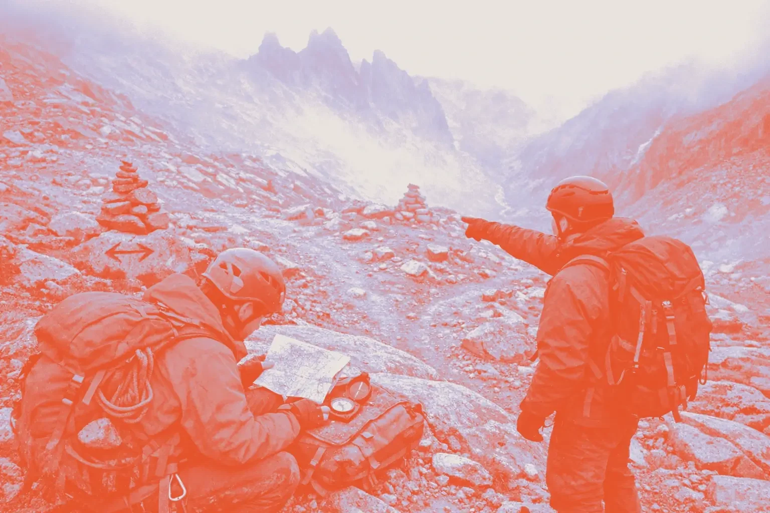 Two hikers with backpacks consult a map near a stack of stones in a misty mountainous area.