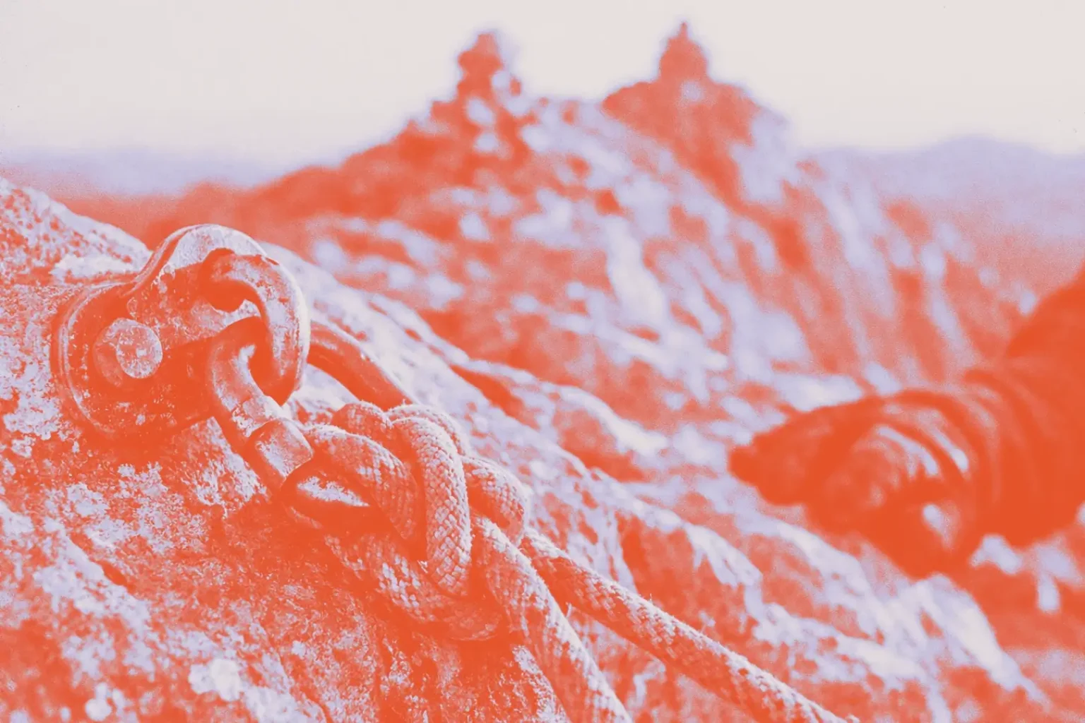 A weathered metal climbing anchor bolted into rock with a knotted rope threaded through it, a blurred mountain ridge in the background, shot in warm editorial tones.