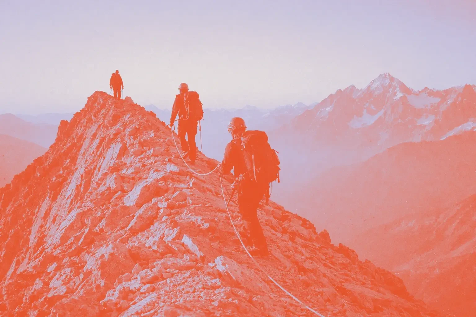Four climbers walk along a rocky mountain ridge with a snowy peak in the background.