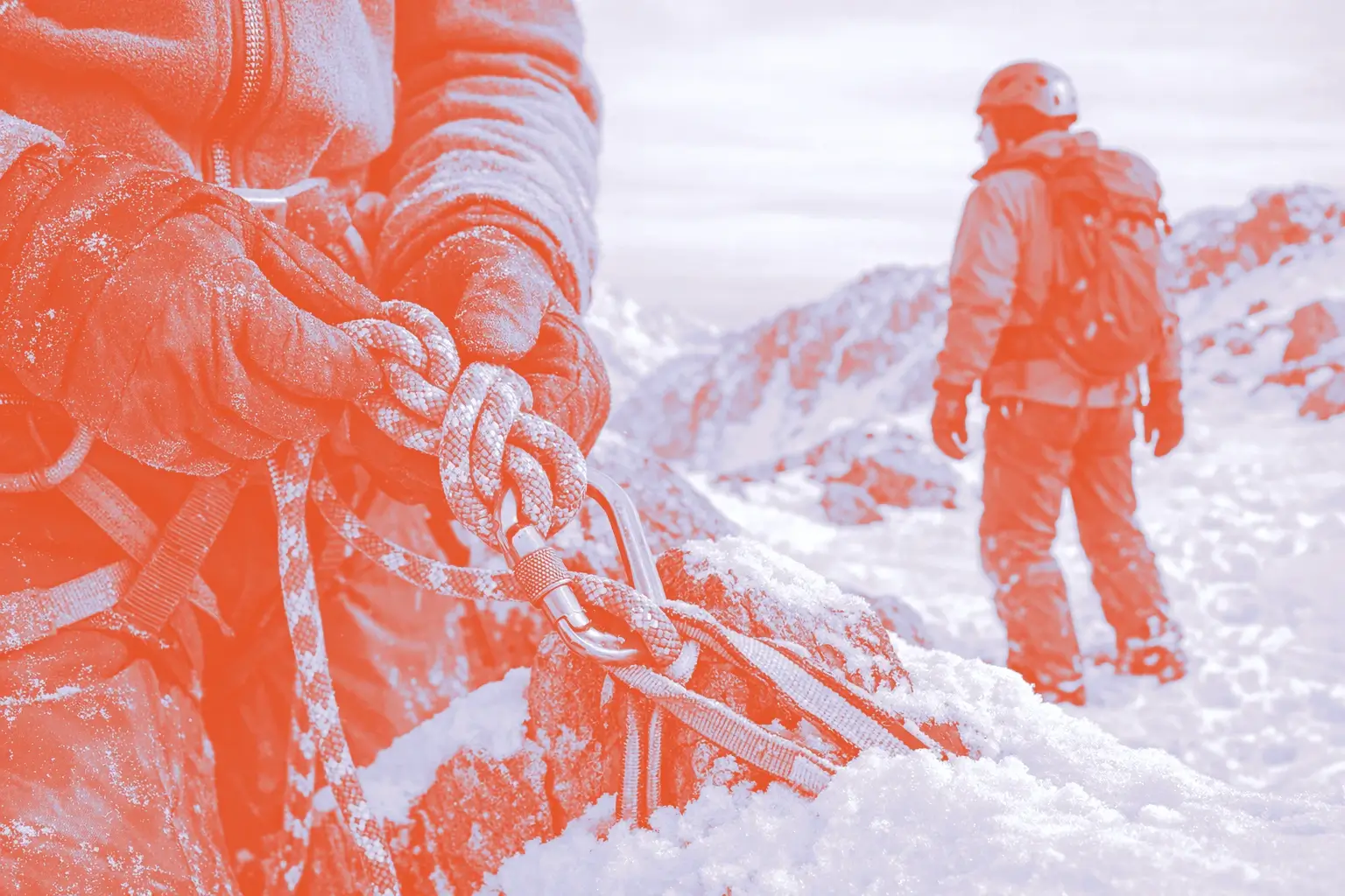 A close-up of a person's hands tying a knot with climbing rope, with a person in red standing in the background on snow-covered rocks.