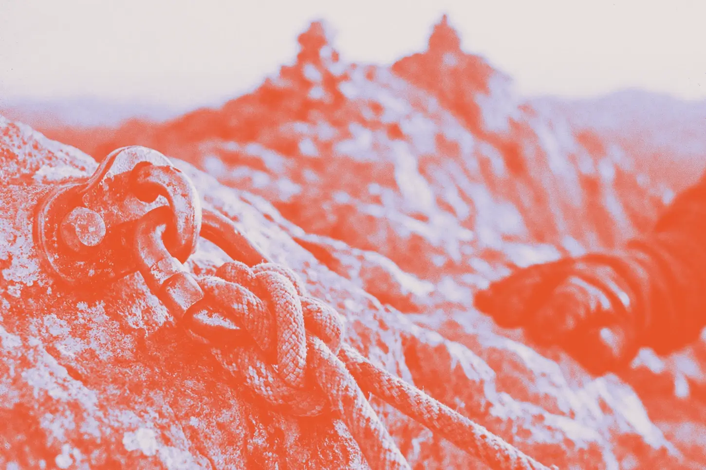 A weathered metal climbing anchor bolted into rock with a knotted rope threaded through it, a blurred mountain ridge in the background, shot in warm editorial tones.