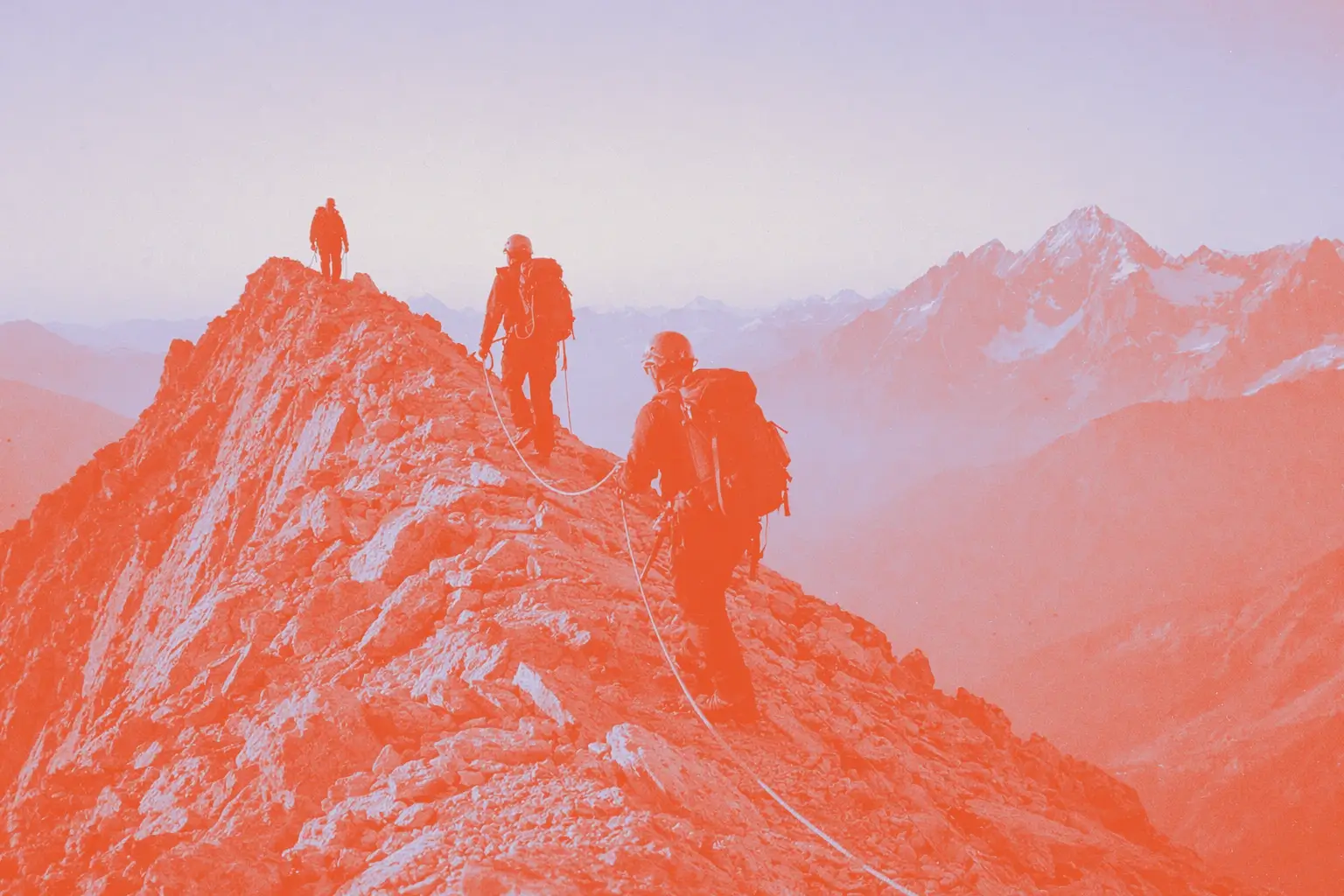 Four climbers walk along a rocky mountain ridge with a snowy peak in the background.